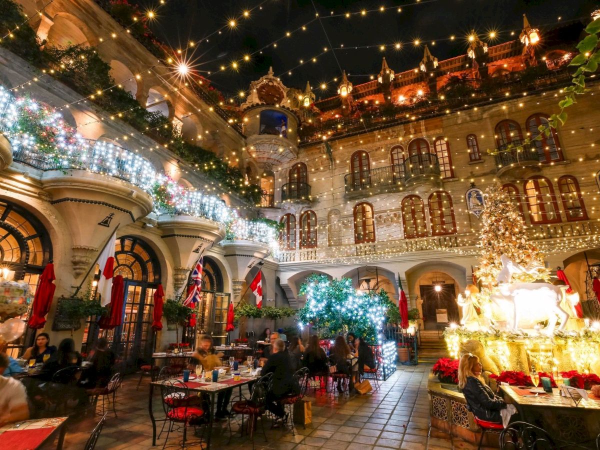 A festive courtyard at night, packed with warm holiday lights, colorful flags, sparkling trees, and a statue display with people dining under the glow.