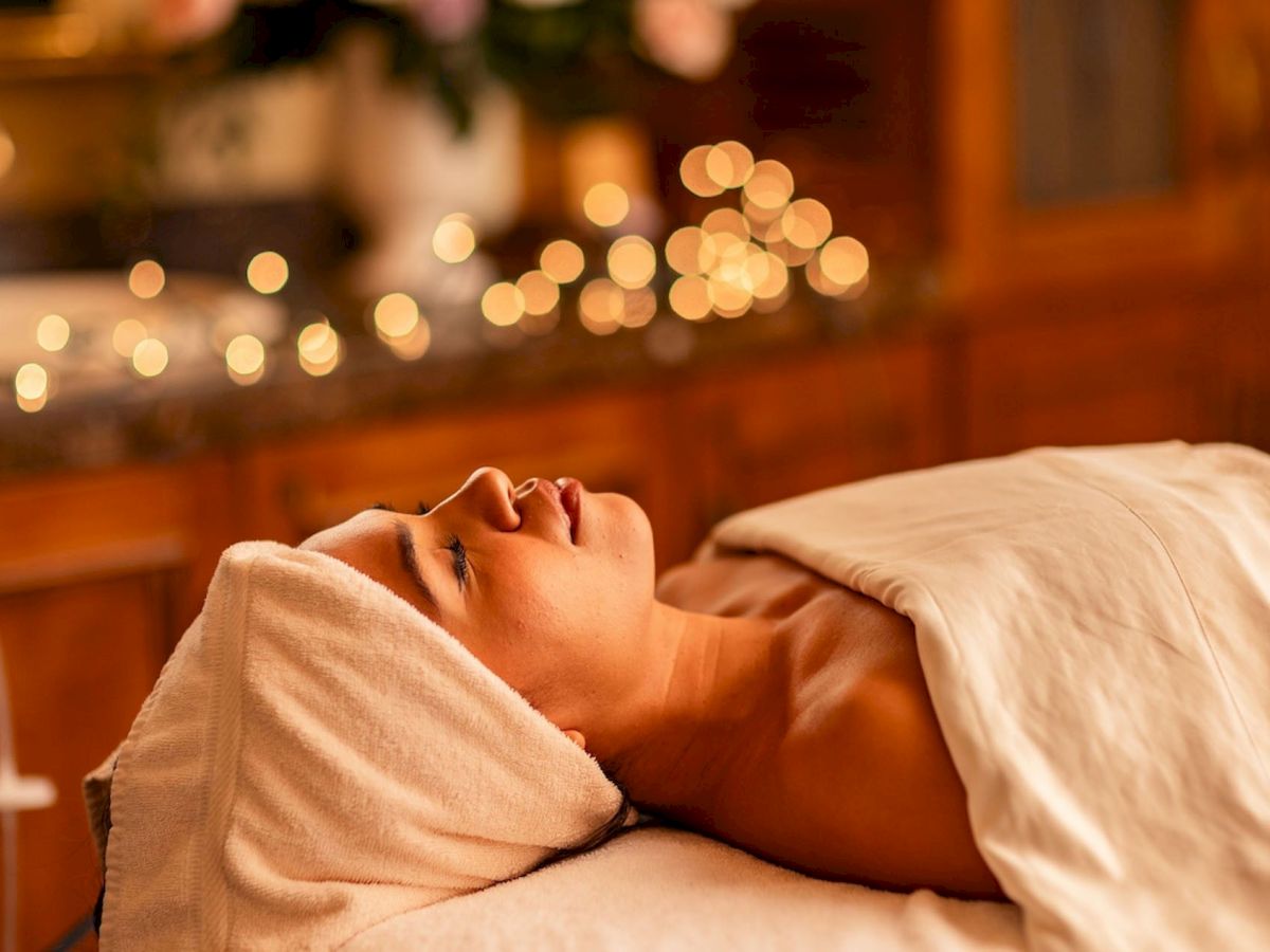 A person lies on a massage table wrapped in a towel in a cozy spa, with warm glowing bokeh lights in the background.