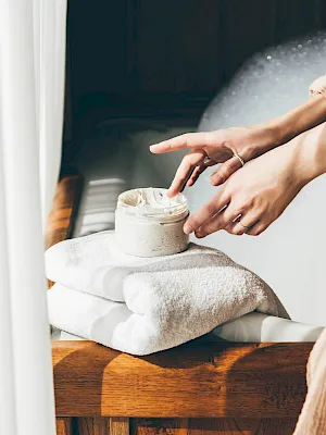 A person’s hands scoop creamy face mask from a jar on a towel, likely for skincare or a spa-style treatment.