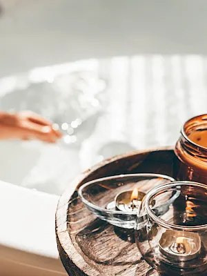 A spa scene with amber essential-oil bottles, small glass cups, and candles beside a hand reaching over a tray of oils.