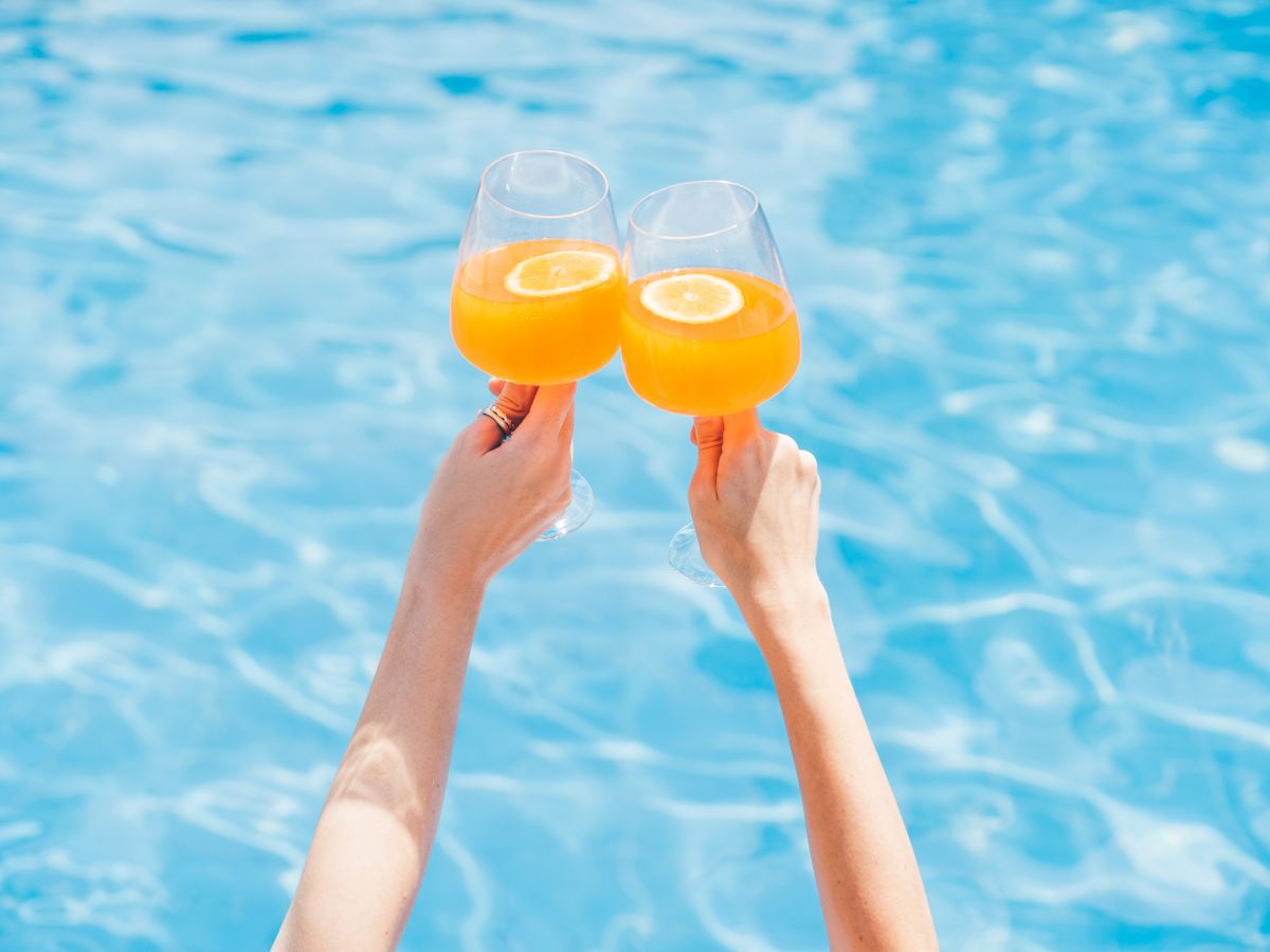 Two hands clink glasses of fresh orange juice over a swimming pool for a sunny toast.