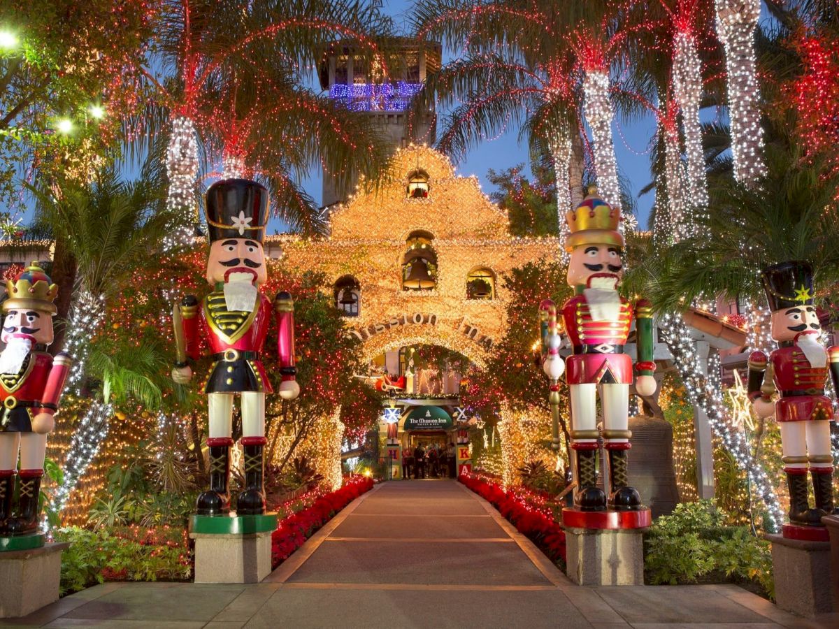 A festive garden walkway lit up with colorful string lights and oversized nutcracker soldiers, with trees wrapped in red, gold, and white glow.