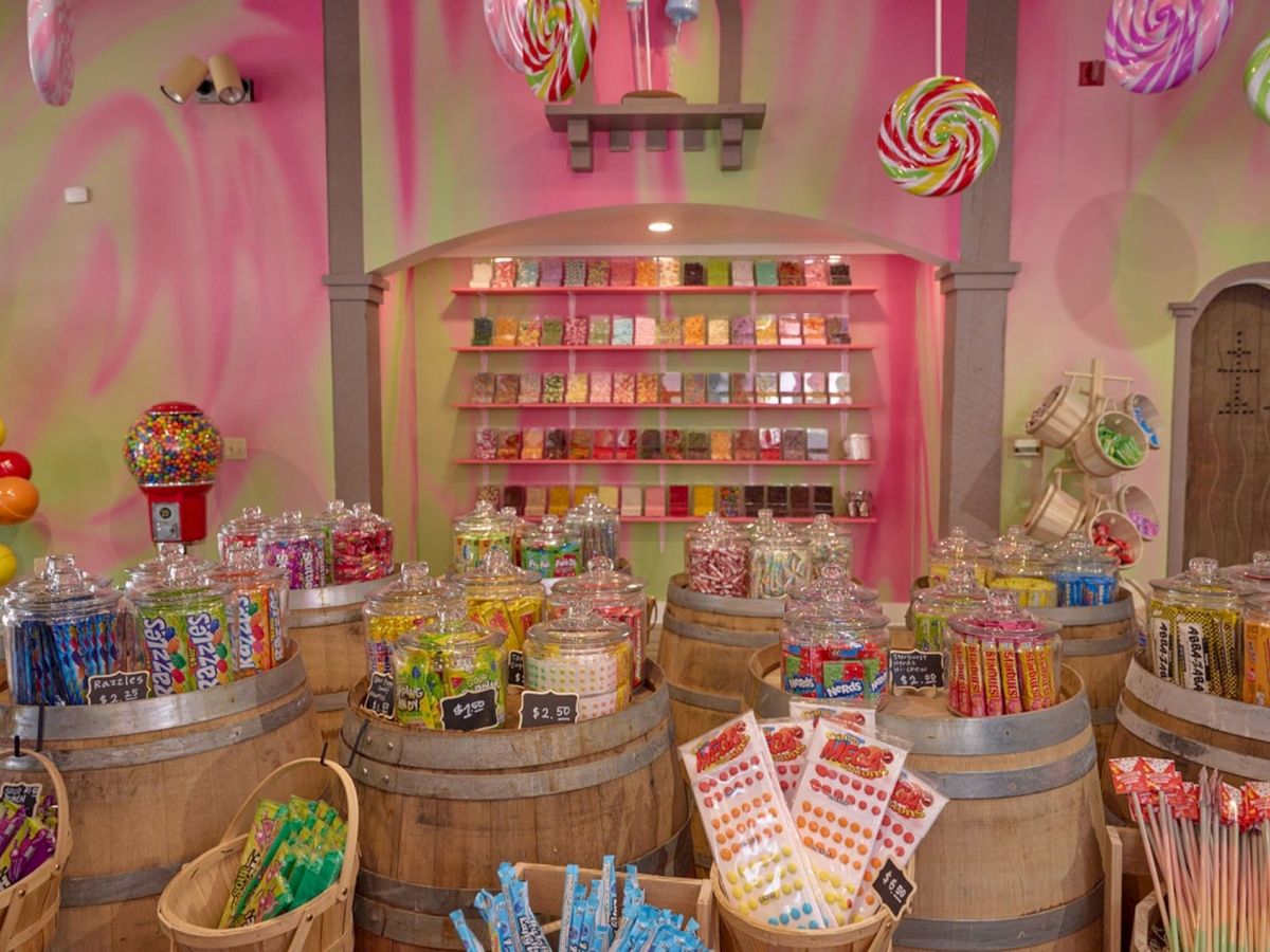 A colorful candy store with jars on barrels, a gumball machine, and swirling lollipop decorations hanging from the ceiling.