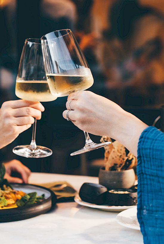 Friends clink wine glasses at a cozy dinner table, toasting with sparkling white wine.