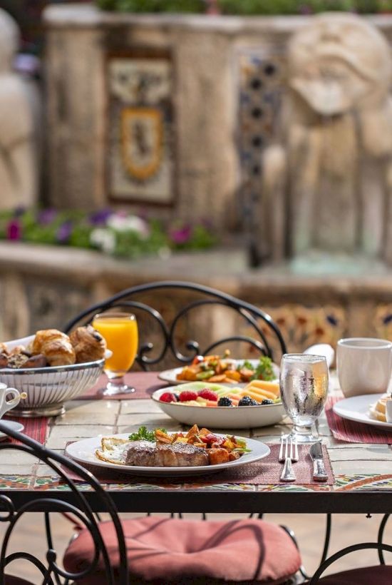 An outdoor patio breakfast table with plates of food and fruit, orange juice, and coffee cups in front of a decorated wall.