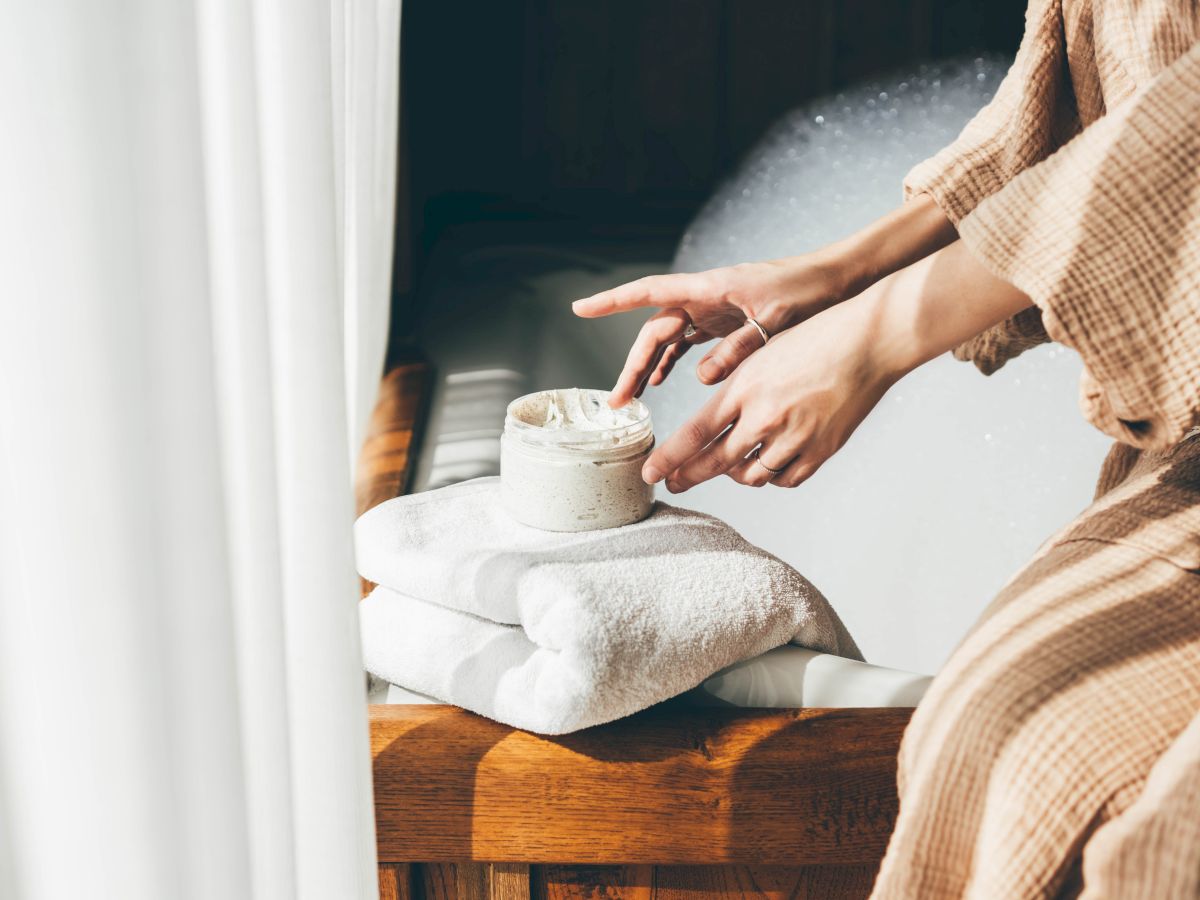 A person scoops cream or scrub from a small jar while resting on a towel by the sink in a bright bathroom.