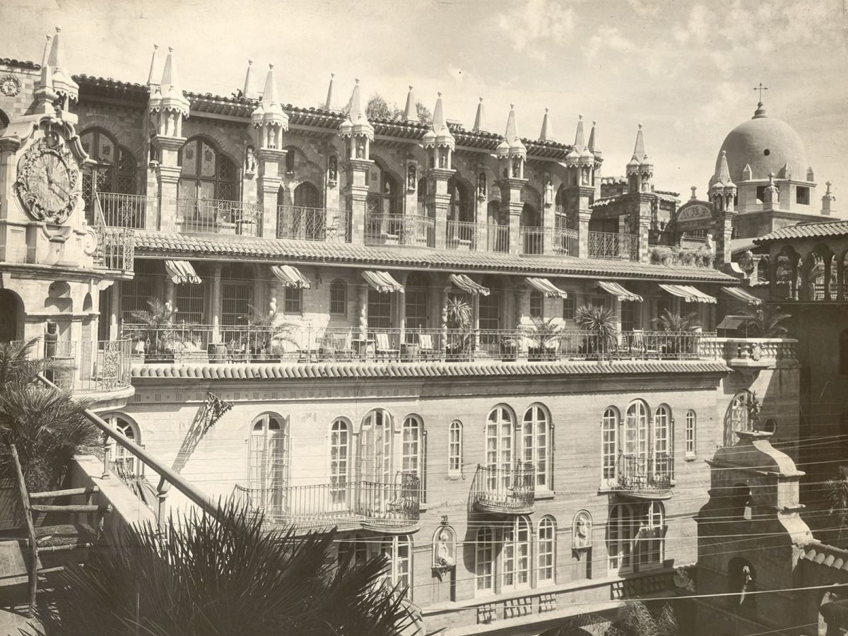 An ornate historic building fa&ccedil;ade with arched windows, balconies, spires, awnings, and a large domed tower in a sunny courtyard.